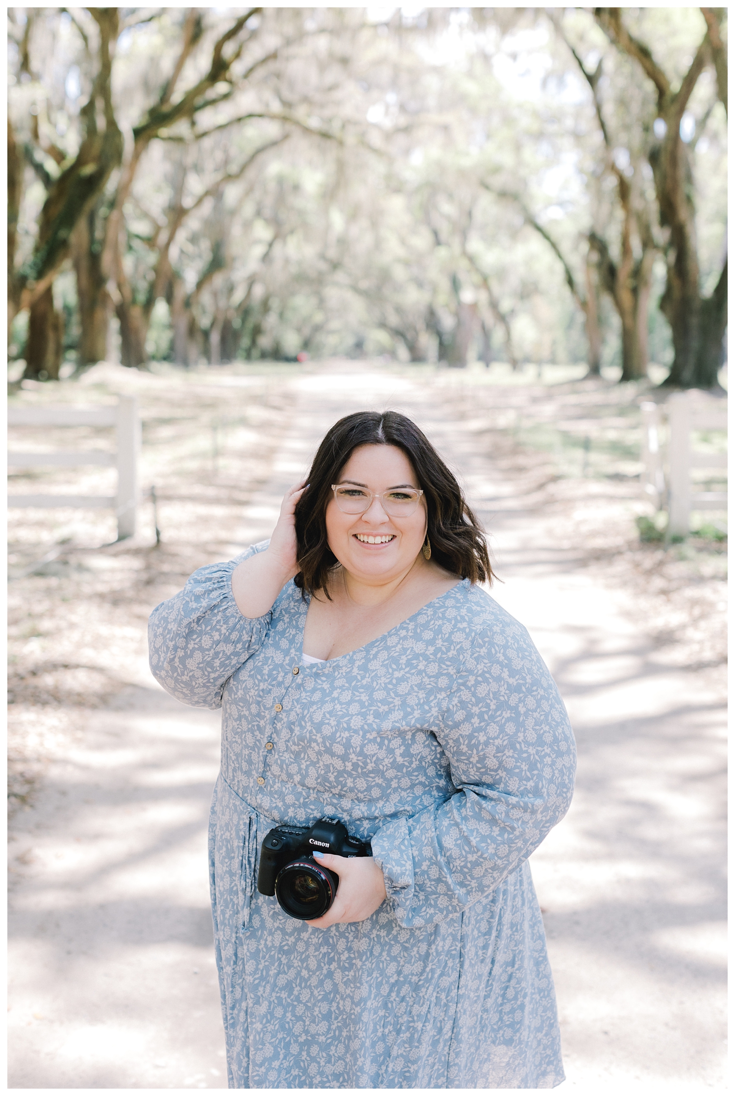 Lexi Alexander in a blue floral dress holding her camera, standing on a sandy lane beneath an avenue of mossy live oaks near Savannah